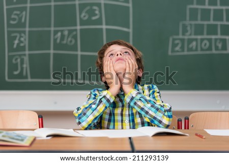 Young schoolboy searching for answers sitting at his desk in the classroom staring up into the air with a thoughtful expression