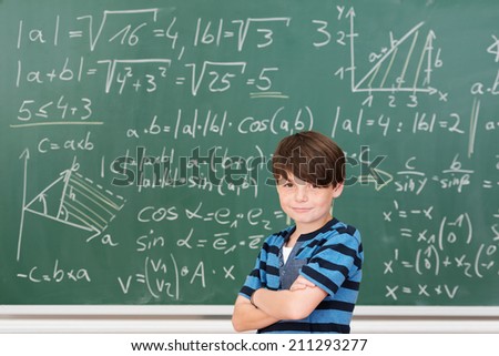 Intelligent kid smiling while posing with folded arms in front of a green blackboard full of mathematical formulas and solved exercises, in the classroom of an elementary school
