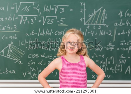 Intelligent little girl child prodigy in class standing confidently in front of a blackboard covered in mathematical equations with her hands on her hips smiling at the camera