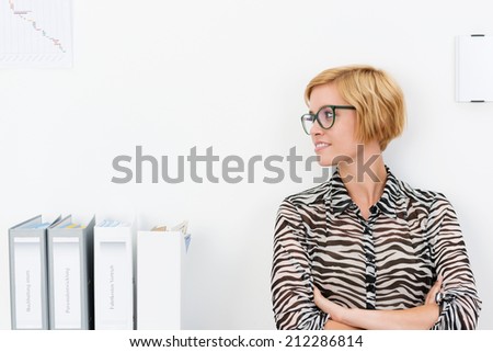 Attractive young woman wearing glasses standing with folded arms in an office looking to the side as she watches something, with copyspace