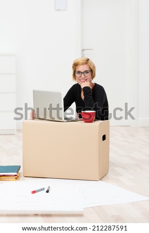 Businesswoman working from home in a new build house sitting on the floor with her laptop balanced on a cardboard packing carton surrounded by large sheets of paper and her coffee