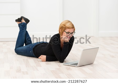 Smiling woman lying on a white parquet floor on her stomach working on her laptop computer smiling as she reads the screen