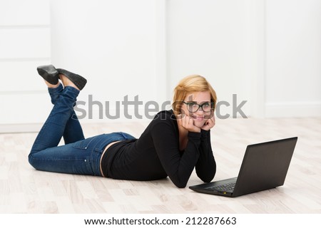 Casual attractive woman working on the floor lying on her stomach with her feet in the air using a laptop computer and smiling at the camera