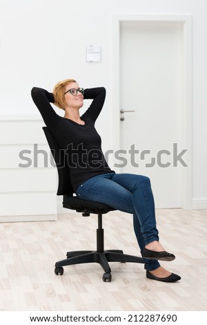 Happy woman relaxing in an office chair in an empty white room leaning back comfortably with her hands clasped behind her head