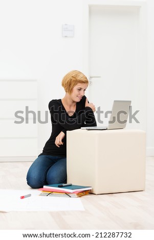 Young woman working on a cardboard box in an empty newly painted white room smiling as she reads her laptop screen after moving house or office
