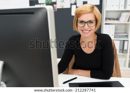 Friendly attractive young businesswoman sitting at her desk in the office smiling at the camera past her computer monitor