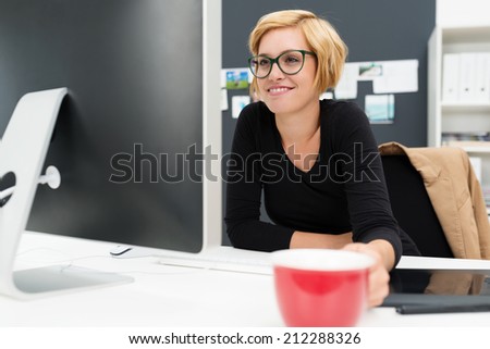 Businesswoman drinking coffee as she works holding a red cup in her hand as she reads information on her computer screen with a relaxed smile