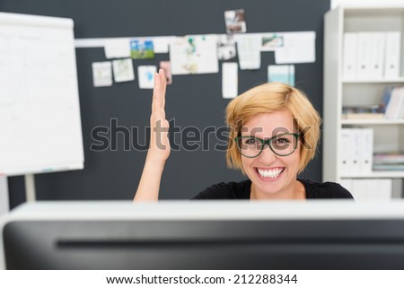 Enthusiastic happy young businesswoman wearing glasses sitting behind her desktop monitor cheering and raising her hand