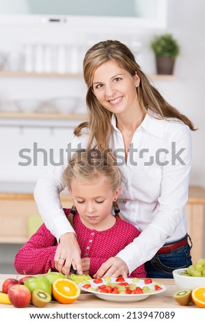 Young mother helping her cute little daughter in the kitchen as they stand together at the counter chopping fresh fruit for a delicious fruit salad