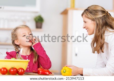 Cute little girl tasting the vegetables as she prepares a meal with her mother in the kitchen chewing on a ripe red cherry tomato with a look of bliss