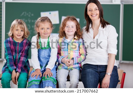 Happy school team of three pretty young girl pupils and their attractive friendly teacher posing together sitting on a table in the classroom