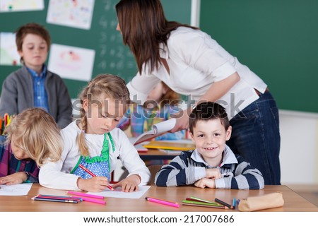 Young female teacher in kindergarten class with a group of diverse young students leaning over to direct a young boy at the back of the classroom