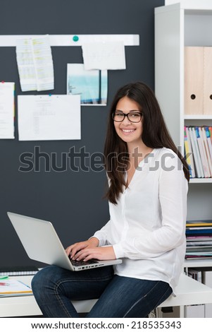 Relaxed young woman working on a laptop balanced on the edge of her desk at the office pausing to smile at the camera