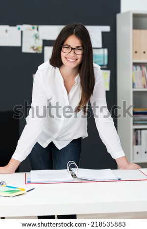 Smiling successful young manageress standing leaning on her desk in the office giving the camera a friendly confident smile