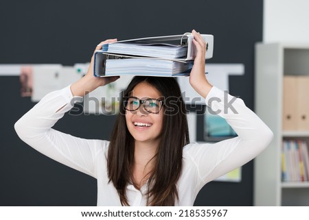 Happy hardworking young businesswoman balancing two large office files o her head as she walks across the office with a lovely smile