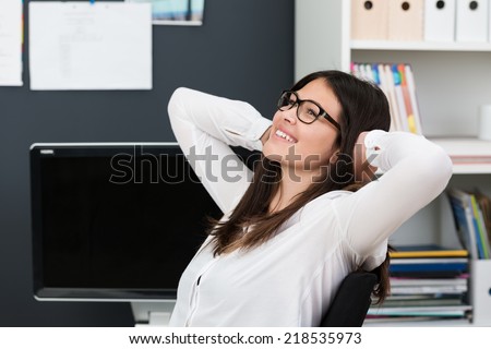 Happy successful young businesswoman wearing glasses relaxing at work leaning back in her chair with a smile of satisfaction and hands clasped behind her head