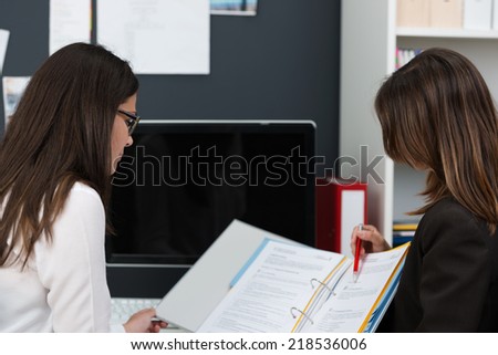 Two young women in the office working on paperwork as they sit together at a desk, over the shoulder view of the file they are working on