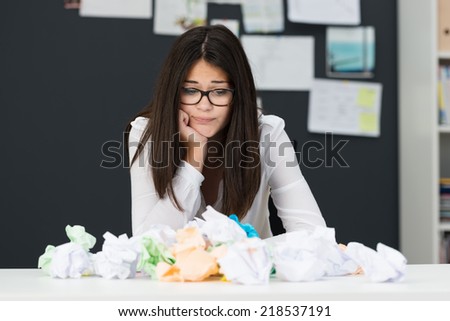 Frustrated young businesswoman with a huge pile of crumpled paper on her desk grimacing as she fails to come up with a solution or creative idea