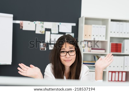 Young businesswoman shrugging her shoulders as she sits at her desk behind a desktop computer to show her ignorance or that she simply does not care