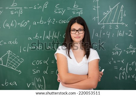 Intelligent young female maths student or teacher wearing glasses standing in front of a chalkboard with mathematical equations with folded arms and a piece of chalk in her hand