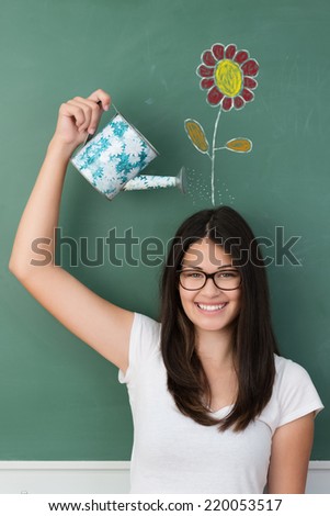 Smiling student watering a colorful flower on her head drawn ion the chalkboard behind in a fun conceptual image