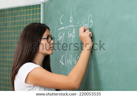 Young female teacher or student wearing glasses standing at the blackboard doing mathematics equations and problems during class in college or university