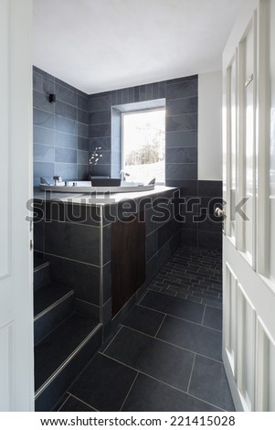 Viewed through a white painted wooden door into a modern tiled bathroom interior with charcoal grey wall and floor tiles and a built in vanity