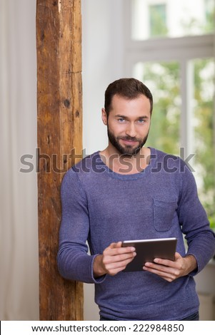 Smiling relaxed bearded man in a casual t-shirt leaning against an old wooden door jamb holding a tablet computer in his hands and smiling at the camera