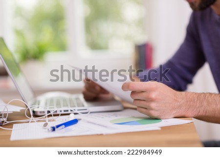 Man sitting working at his desk reading a handheld document as he works on a report and analysis, close up view of his hands and the desk