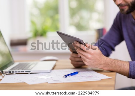 Man working on a handheld tablet computer at his desk in the office reading information on the screen, close up view