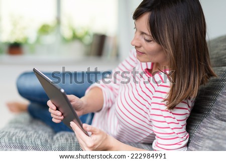 Young woman in casual clothes relaxing at home on a grey sofa reading her tablet computer