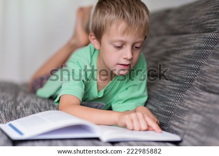 Small boy lying on a sofa reading a big book concentrating carefully following the text with his finger