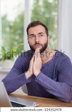 Businessman sitting thinking at his desk with a serious expression and hands clasped as though in prayer staring into the air