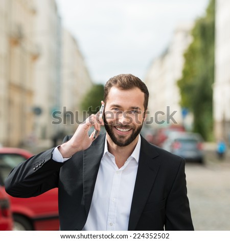 Happy attractive businessman on his mobile phone chatting as he walks along an urban street looking at the camera with a beaming smile