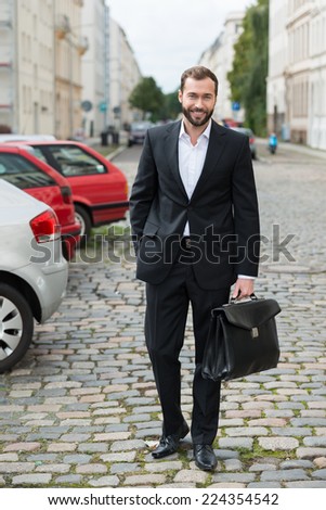 Stylish businessman walking to work across a cobbled parking lot carrying his briefcase smiling at the camera