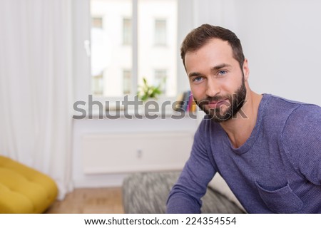 Attractive bearded man with a friendly smile sitting on a sofa in his apartment looking at the camera