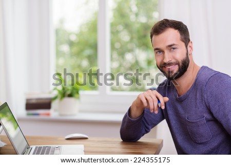 Good-looking bearded man in a home office sitting at a table with his laptop computer turning to smile at the camera