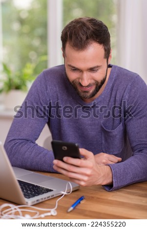 Man smiling as he reads a message on his mobile while working at his desk in the office at home