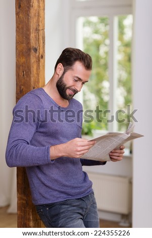Relaxed man standing reading a newspaper while leaning against an old wooden door jamb at home smiling as he reads the news