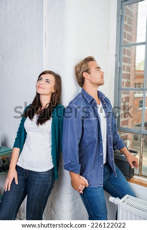 Young couple standing daydreaming in their apartment leaning back to back against the wall looking dreamily up into the air