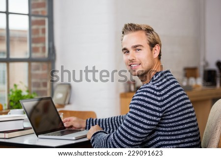 Handsome young man in casual clothes working from home on his laptop computer turning to smile at the camera