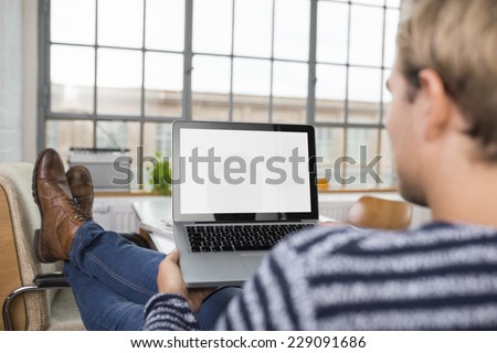 Over the shoulder view of a man with his feet up working on a laptop at home with a blank screen visible to the viewer