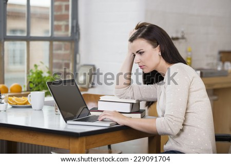 Harassed young woman working on a laptop resting her arm on a pile of books as she frowns at the screen while sitting at a table in her apartment