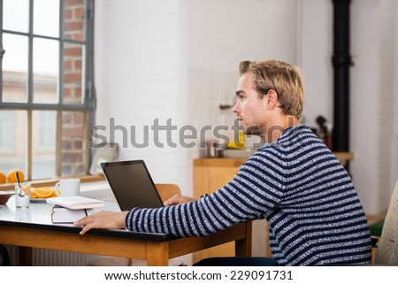 Serious young man working at home on his laptop computer sitting staring out of a window with a thoughtful expression