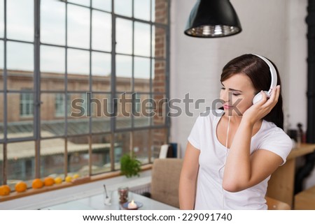 Young woman enjoying her music at home standing in the kitchen holding the headphones to her ear with a smile of bliss