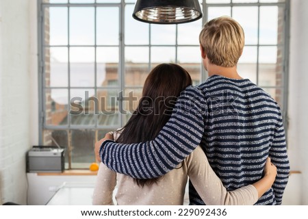 Young couple in love standing arm in arm with their backs to the camera looking through their apartment window at urban buildings
