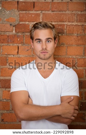 Gorgeous Young Blond Man in White Shirt Crossing Arms in Front on Bricks Wall Background. Looking at Camera.