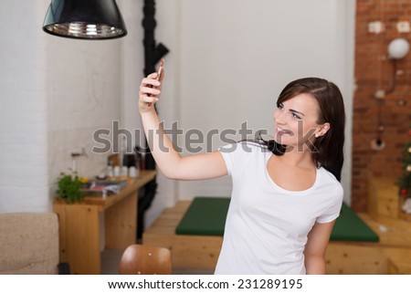 Attractive casual young woman posing for a selfie on her mobile phone indoors at home smiling at the camera