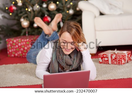 Young woman relaxing with her laptop in front of a Xmas tree lying barefoot on the floor surrounded by Christmas gifts