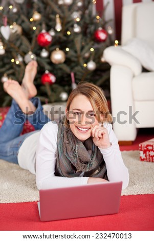 Pretty Smiling Woman Lying in Prone on the Floor, with Laptop Computer, Near Christmas Tree Decoration While Looking at the Camera.
